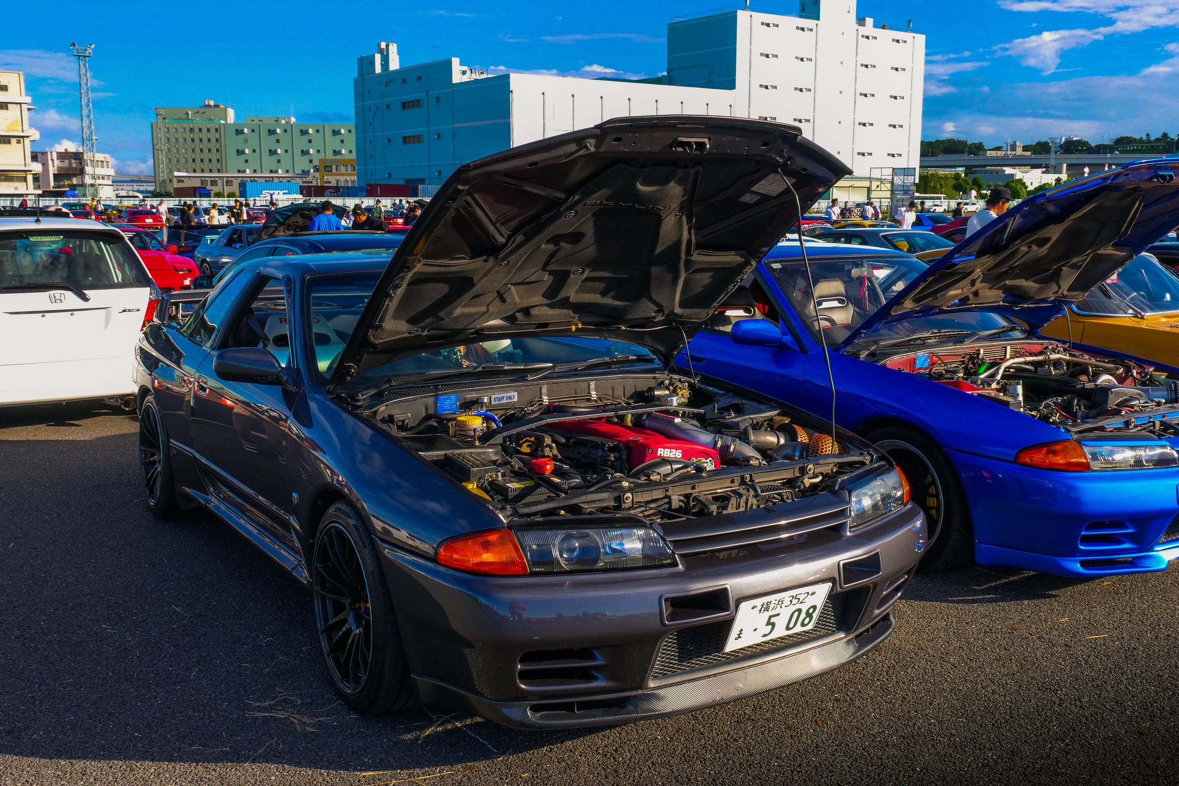Nissan Skyline GT-R R32 grise, capot ouvert sur son six-cylindres RB26 au cache rouge, à côté d'une R32 bleue, lors d'un rassemblement de voitures au Japon.