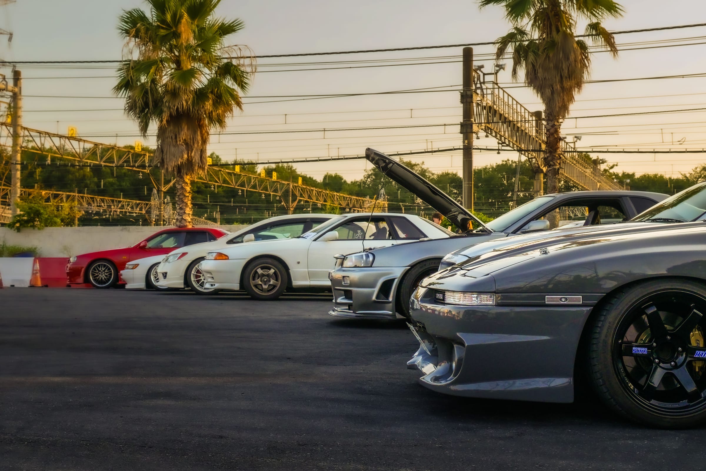 Sportives japonaises des années 90 alignées au coucher du soleil lors d'un rassemblement à Tokyo : un coupé gris au premier plan, une Nissan Skyline R34 grise capot ouvert, une Skyline R32 blanche.