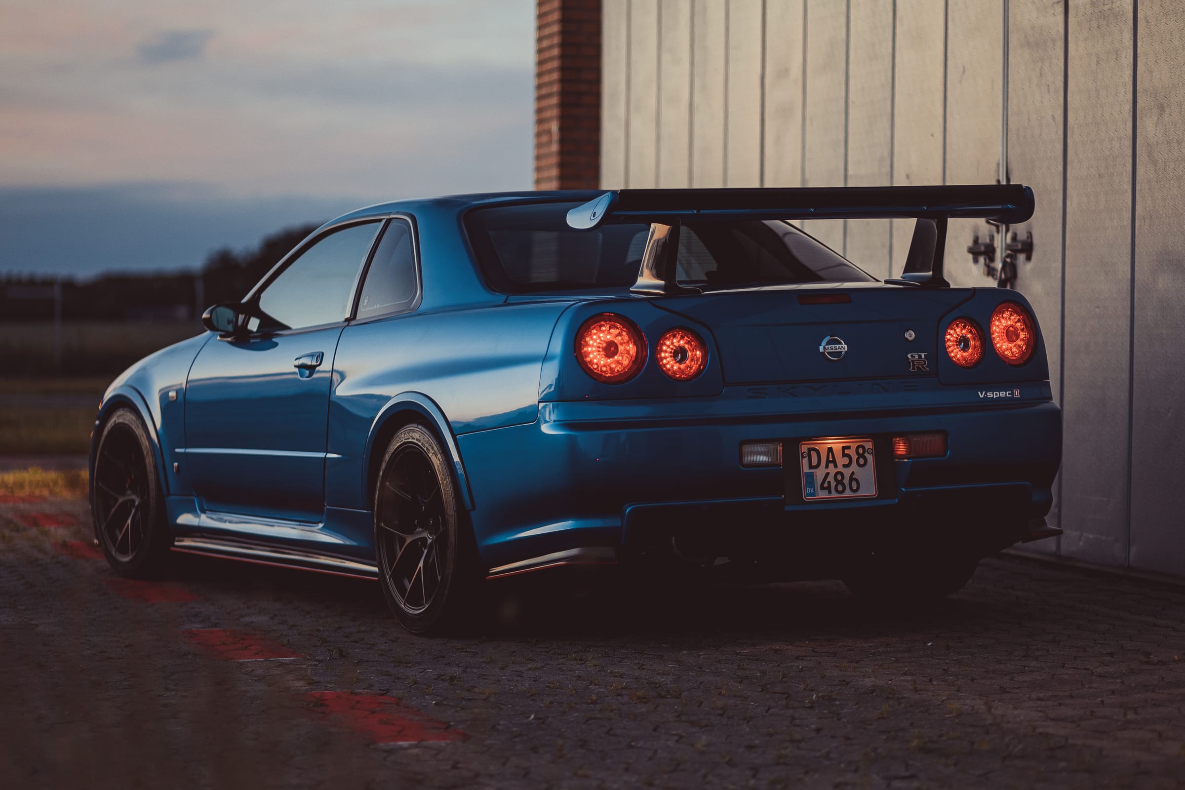 Nissan Skyline GT-R R34 V-spec II bleue vue de trois quarts arrière au crépuscule, ses quatre feux ronds allumés.