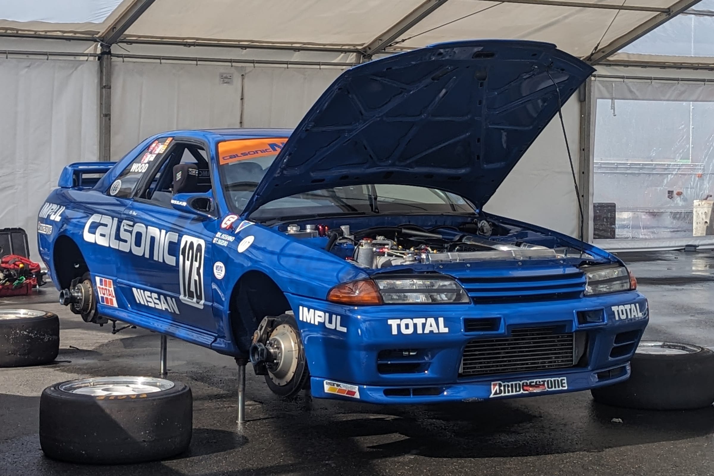 Nissan Skyline GT-R R32 de course bleue en livrée Calsonic, capot ouvert et roues démontées, sur chandelles dans un paddock.
