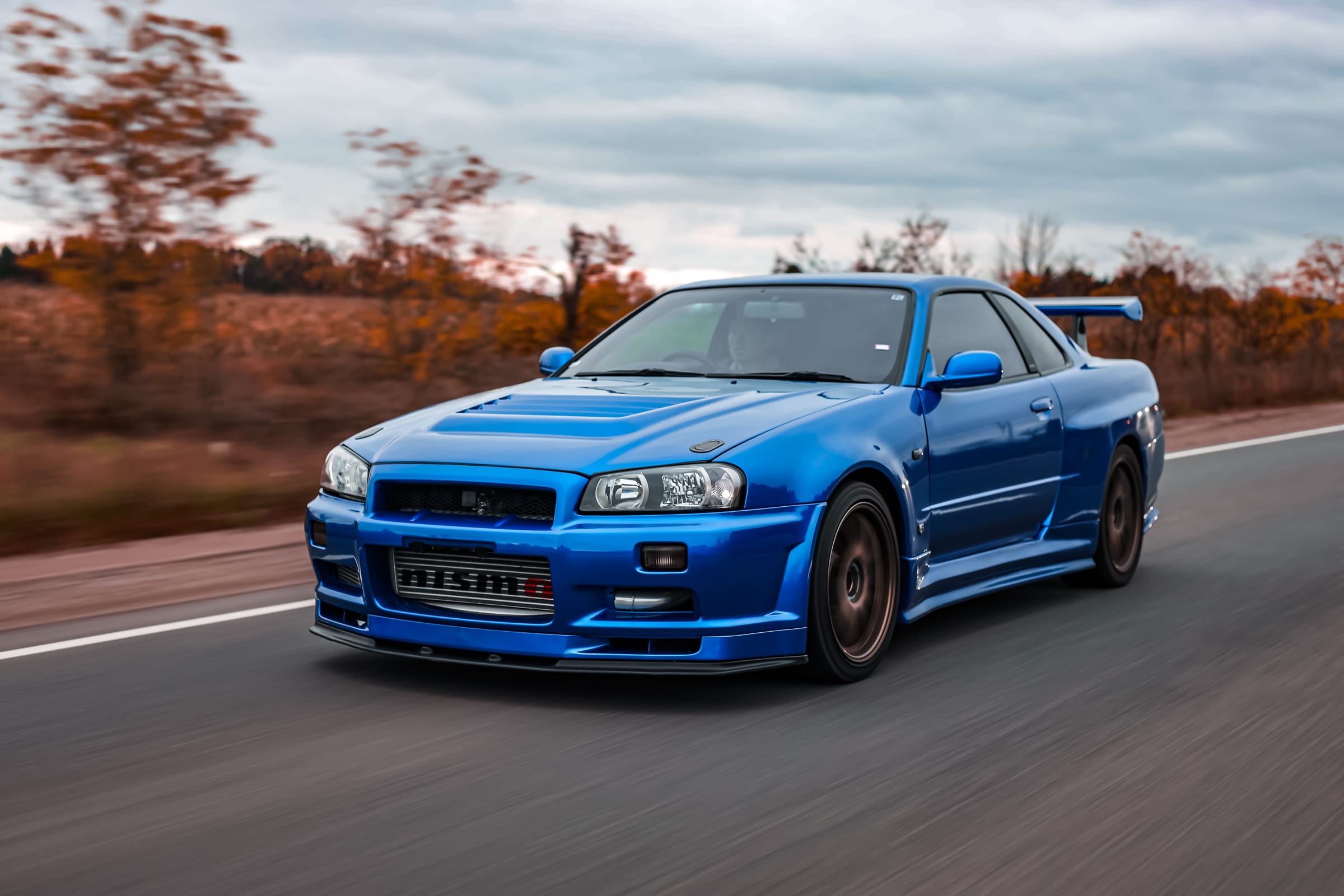 Nissan Skyline GT-R R34 bleue en mouvement sur une route bordée d'arbres aux couleurs d'automne.